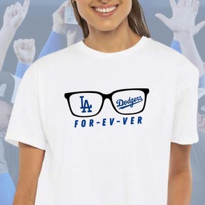 May include: White t-shirt featuring a black glasses graphic with the Los Angeles Dodgers logo and the text "FOR-EV-VER" in blue. The shirt has a crew neck and short sleeves. The image also includes a "Bleed Blue Designs" logo.