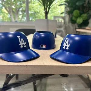 May include: Three blue plastic baseball caps with the LA logo. One cap has a World Series Champions patch. The caps are for holding drinks.