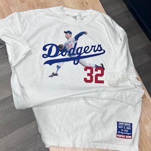 May include: White t-shirt with a blue and white "Dodgers" logo and a photo of a baseball player wearing a blue and white uniform with the number "32" on the front. The text "SANDY KOUFAX SEPT 9, 1965 VS CUBS LOS ANGELES DODGERS PERFECT GAME" is printed on a white tag attached to the bottom of the shirt.