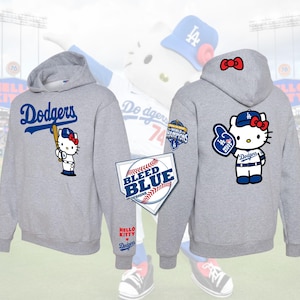 May include: Gray hooded sweatshirts with the word "Dodgers" in blue script. One sweatshirt features a Hello Kitty character holding a baseball bat, and the other has Hello Kitty with a foam finger. The sweatshirts have the text "BLEED BLUE DESIGNS" and "HELLO KITTY Dodgers".