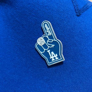 Peut inclure: Épinglette bleue et blanche en forme de doigt de mousse, avec les mots "Los Angeles" et la lettre "A". L'épingle est posée sur un tissu bleu. Accessoire de fan.