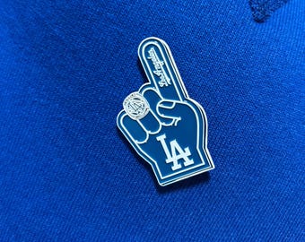 May include: A blue and white pin shaped like a foam finger, with the words "Los Angeles" and the letter "A" visible. The pin is on a blue fabric background. The pin is a fan accessory.