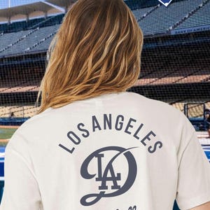 Puede incluir: Top corto blanco con el texto "City Connect Crop" y un logotipo y texto azul marino de los Dodgers de Los Ángeles en la espalda. Tema de béisbol.