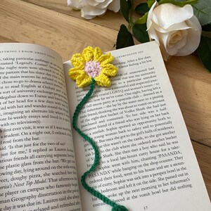 May include: A yellow crocheted flower bookmark with a green stem and leaves. The bookmark is on top of an open book with text about a university exchange programme.