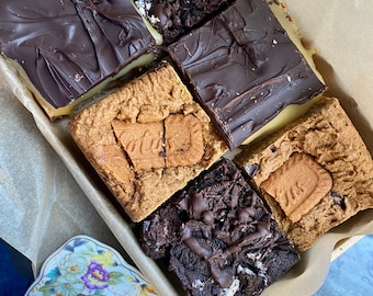 May include: A variety of homemade brownies in a brown paper-lined tray. The brownies have different toppings, including chocolate ganache, cookie pieces, and a cookie. A teacup and saucer with a floral design are in the foreground.