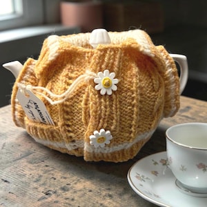 May include: A white ceramic teapot with a mustard yellow knitted tea cozy. The cozy has a cable knit design, white trim, and daisy-shaped buttons. A teacup and saucer with floral patterns are on the right.