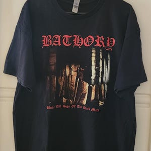 Op de afbeelding: Zwart t-shirt met de bandnaam "BATHORY" in rode gotische letters. Het shirt heeft een afbeelding van een donkere, gotische scène en de tekst "Under The Sign Of The Black Mark".