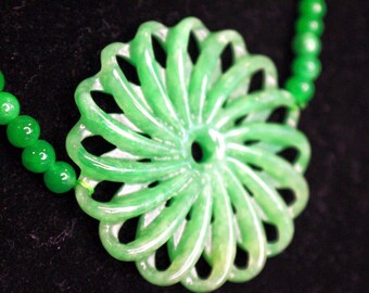 May include: A green jade pendant necklace with a carved flower design. The pendant is a large, intricate flower with a hole in the center. The necklace is made of small, round jade beads.