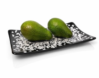 May include: Two green avocados rest on a black rectangular plate with a white speckled pattern. The plate has slightly curved edges and sits on a white surface. The avocados are smooth and vibrant green.