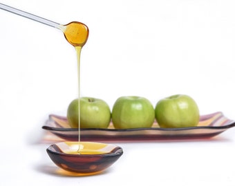 May include: A glass bowl filled with honey, with a spoon dripping honey over the side. Three green apples are on a striped plate in the background.