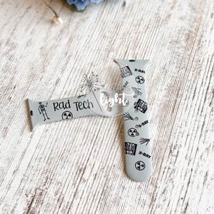 May include: A light gray silicone watch band featuring black printed graphics of skeletons, X-ray images, and radiation warning symbols. The text "Rad Tech" is printed on one side of the band, which includes multiple adjustment holes and a connector.