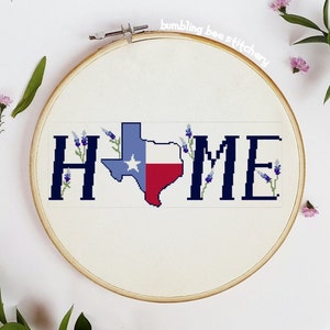 May include: Cross-stitch embroidery in a wooden hoop. The design features the word "HOME" with the Texas state outline in the center, colored in the state flag colors. Lavender sprigs are placed around the letters and state outline.