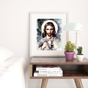 Puede incluir: Una pintura de acuarela de Jes&uacute;s sosteniendo un c&aacute;liz y una hostia. La pintura est&aacute; enmarcada en un marco blanco y est&aacute; colgada en una pared sobre una mesa de madera con libros y una l&aacute;mpara.