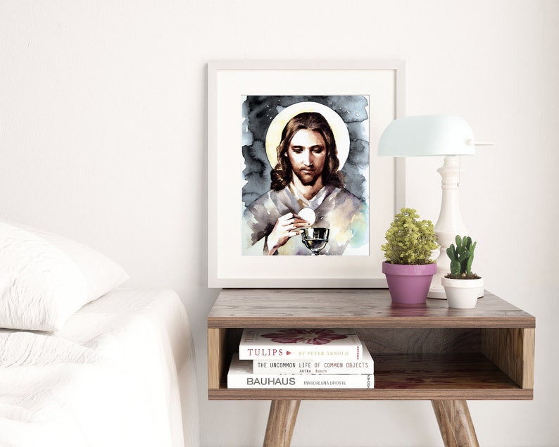 Puede incluir: Una pintura de acuarela de Jes&uacute;s sosteniendo un c&aacute;liz y una hostia. La pintura est&aacute; enmarcada en un marco blanco y est&aacute; colgada en una pared sobre una mesa de madera con libros y una l&aacute;mpara.