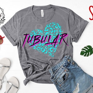 Puede incluir: Una camiseta gris con un diseño de corazón azul y rosa que dice "TUBULAR" en una fuente en negrita. La camiseta está sobre una superficie blanca con un par de sandalias plateadas, un llavero de hoja verde y un par de gafas de sol rojas.