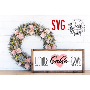 Puede incluir: Un letrero de madera con el texto "LITTLE babe CAVE" en letras negras. Un corazón rosa con un contorno blanco se encuentra entre las palabras "babe" y "CAVE". El letrero está sentado sobre una superficie de madera frente a una corona de flores rosas y hojas verdes.