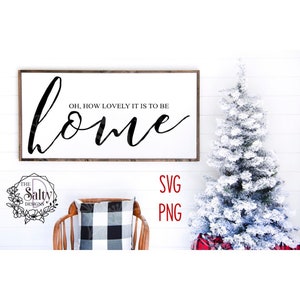 Puede incluir: Un cartel blanco con un marco de madera marrón dice "Oh, how lovely it is to be home" en escritura cursiva negra. El cartel está colgado en una pared blanca con un árbol de Navidad blanco y un cojín a cuadros blanco y negro en una silla de madera en el fondo.