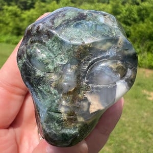 May include: A carved stone sculpture of an alien head. The sculpture features a detailed face with large, oval eyes and a subtle smile. The stone is a mix of green, brown, and white, with a polished finish. The sculpture is a unique decorative item.