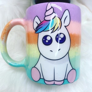 May include: A white ceramic mug with a rainbow stripe design and a cartoon unicorn with a rainbow mane and horn. The unicorn has big blue eyes and is sitting down.