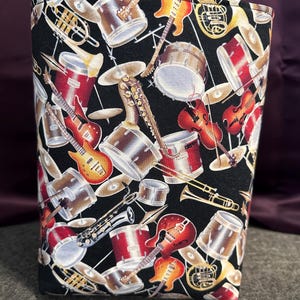 May include: Black fabric with a repeating pattern of musical instruments, including drums, guitars, saxophones, trumpets, and violins. The instruments are in shades of red, yellow, and gold.