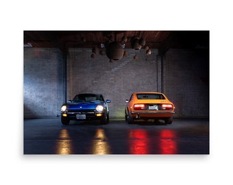 May include: Two classic sports cars, one blue and one orange, are parked side-by-side in a garage. The blue car faces forward, while the orange car is seen from the rear. The garage has a brick wall and hanging lights.