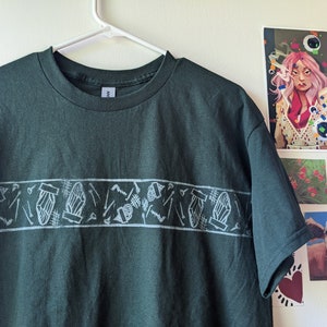 May include: Dark green t-shirt with a white graphic band across the chest. The band features a repeating pattern of stylized objects. The shirt is hanging on a white hanger against a white wall with artwork.