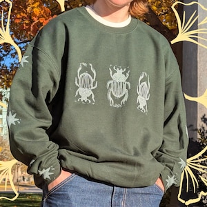 May include: Dark green sweatshirt with a white crew neck and a light green insect graphic. The graphic features three stylized beetles. The sleeves have star designs. The person is wearing blue jeans.