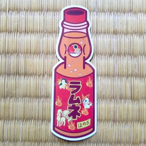 Puede incluir: Una pegatina con una ilustración de dibujos animados de una botella de refresco Ramune japonesa. La botella es naranja con una etiqueta roja y texto japonés. El diseño incluye personajes de dibujos animados y llamas.