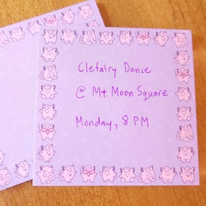 May include: Two lavender-colored square cards with a border of pink cartoon characters. One card has handwritten text: "Clefairy Dance @ Mt. Moon Square Monday, 8 PM". The other card is blank. The cards are on a wooden surface.