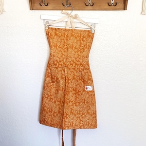 以下が含まれることがあります： 白いハンガーに吊るされた、花柄のオレンジ色のエプロン。エプロンには2つのポケットと右ポケットに小さなラベルが付いています。エプロンは首の周りにクリーム色のネクタイとウエストタイが付いています。エプロンは白い壁に掛かっています。