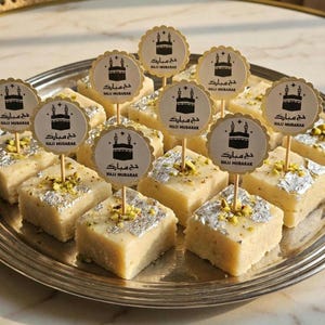 May include: A silver platter displays square desserts, garnished with silver leaf and pistachios. Each dessert has a pick with a black Kaaba illustration and the text "HAJJ MUBARAK". The treats are arranged in rows. The image has a warm, inviting tone.