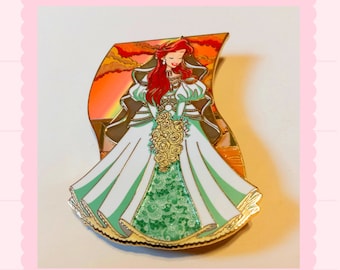 May include: A gold-tone enamel pin featuring Ariel from The Little Mermaid dressed in a white wedding gown with a green and white floral pattern. She is holding a bouquet of yellow flowers and has red hair. The background is a sunset scene.