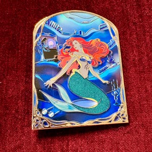 May include: A rectangular pin featuring a mermaid with red hair and a teal tail, set against a blue background. The pin has a gold border and depicts an underwater scene with a castle and other details.