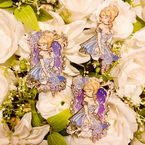 May include: Enamel pins featuring a blonde woman in a blue dress, set against a purple starry background within a gold frame. The pins are adorned with blue and purple floral accents, set on a bed of white flowers.
