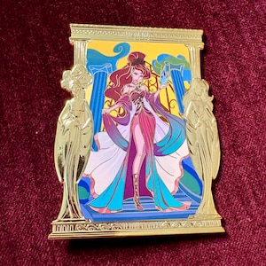 May include: A gold-colored enamel pin featuring a woman in a flowing gown, set against a backdrop of columns and a yellow background. The woman has red hair and is holding a blue object. The pin has a decorative border and two gold statues on either side.