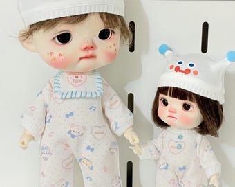 May include: Two dolls wearing matching white pajamas with a pattern of cats, hearts, and musical notes. Each doll wears a white hat with a cartoon face and blue pom-poms. One doll has a bib. They stand on a blue rug.
