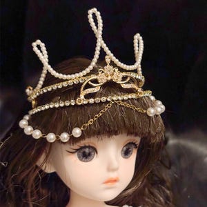 May include: A gold-toned tiara with faux pearls and rhinestones. The crown features a central floral design and dangling chain accents. The tiara is worn by a doll with brown hair and gray eyes, creating a delicate and elegant aesthetic.
