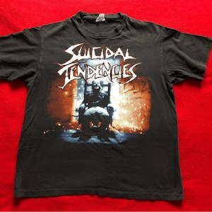 May include: Black t-shirt with a graphic print of the band "Suicidal Tendencies" in white lettering. The graphic features a person sitting on a throne in a dark room with a spotlight shining on them.