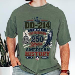 May include: An olive green t-shirt featuring a graphic with a bald eagle, two military dog tags, and the text: My DD-214 IS MY CONTRIBUTION TO 250 Years OF AMERICAN HISTORY 1776 2026. The design includes a shield emblem and stars in red, white, and blue.