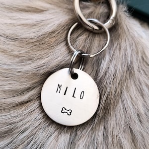 Peut inclure: Une médaille de chien en argent avec le nom "Milo" gravé dessus, avec un petit symbole d'os en dessous du nom. La médaille est attachée à un porte-clés en argent.