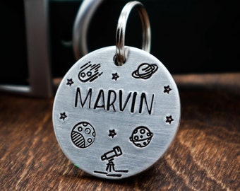 May include: Silver dog tag with a space theme. It has a black engraved design of planets, stars, and a telescope. The name "MARVIN" is engraved in all caps.