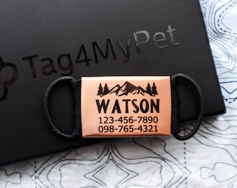 Peut inclure: Une étiquette de chien personnalisée en cuivre avec une scène de montagne et le nom "WATSON" gravé dessus. L'étiquette comprend également deux numéros de téléphone : 123-456-7890 et 098-765-4321. L'étiquette est attachée à une sangle noire.