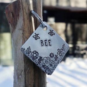 May include: A silver diamond-shaped tag with a floral design and the word "BEE" engraved on it. The tag is attached to a wooden post with a metal ring.