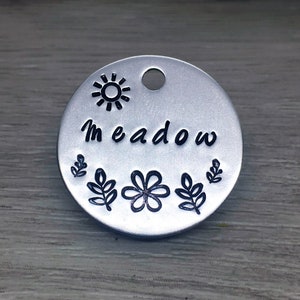 May include: Silver round tag with a sun, leaves, and a flower design. The tag says "Meadow" in black lettering.