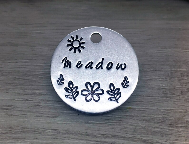 May include: Silver round tag with a sun, leaves, and a flower design. The tag says "Meadow" in black lettering.