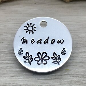 May include: Silver round tag with the word "Meadow" engraved in black lettering. The tag is decorated with a sun, a flower, and leaves, all in black.