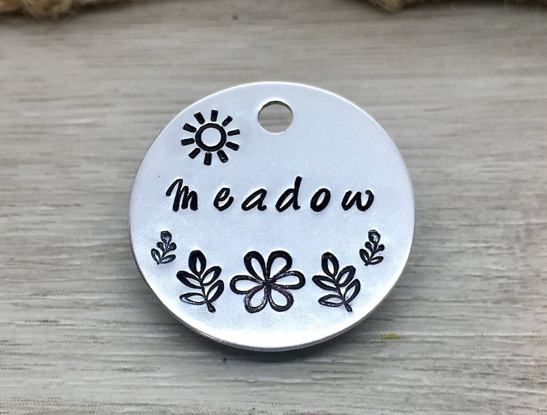 May include: Silver round tag with the word "Meadow" engraved in black lettering. The tag is decorated with a sun, a flower, and leaves, all in black.