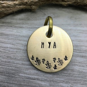 以下が含まれることがあります： 「MYA」と刻まれた丸い真鍮製の犬用タグ。タグは小さな葉のデザインの縁取りで飾られています。