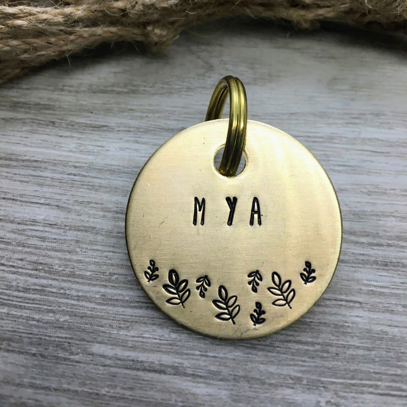 以下が含まれることがあります： 「MYA」と刻まれた丸い真鍮製の犬用タグ。タグは小さな葉のデザインの縁取りで飾られています。