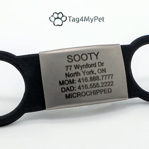 Op de afbeelding: Een zilveren hondenpenning met zwarte rubberen ringen. De penning is gegraveerd met de naam "SOOTY", een adres, telefoonnummers en de tekst "MICROCHIPPED".